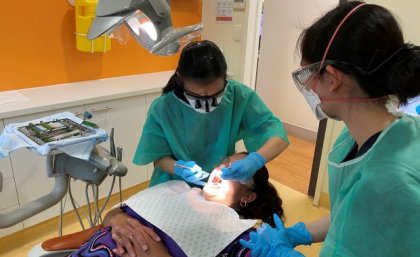 A patient lies on a reclining chair in a dental clinic as two student dentists in scrubs treat her.